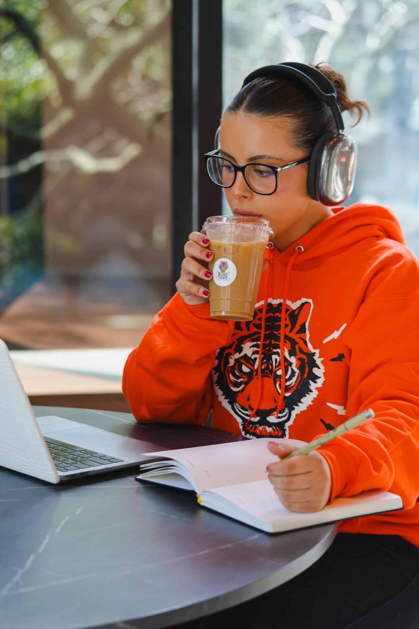 Person in orange hoodie with tiger design drinking coffee and using laptop outdoors