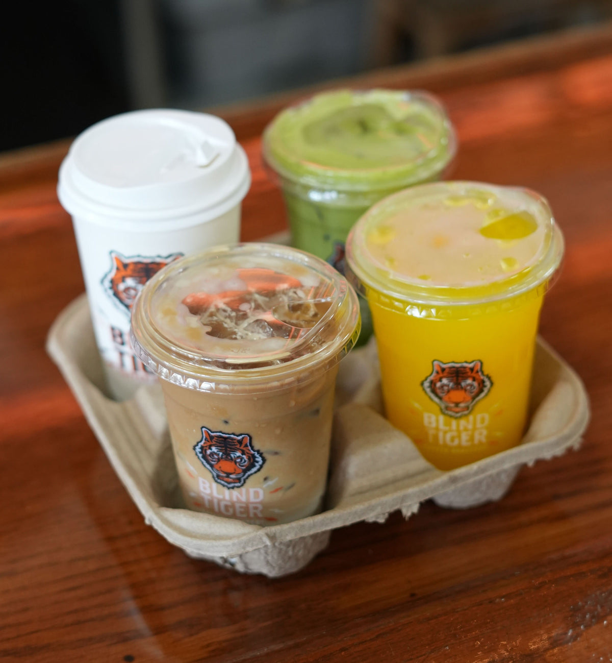 Three branded plastic takeaway cups in a cardboard cup holder on a wooden table. One cup is white with a lid, one is yellow, and one is green; all have the same bear logo and the text 'BIRCH TICKER' visible.