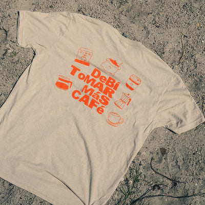 A beige t-shirt laid flat on sandy ground with sparse green grass. The shirt features a bold orange graphic print reading 'Debi TOMAR MAS CAFE' surrounded by coffee cup and bean illustrations.