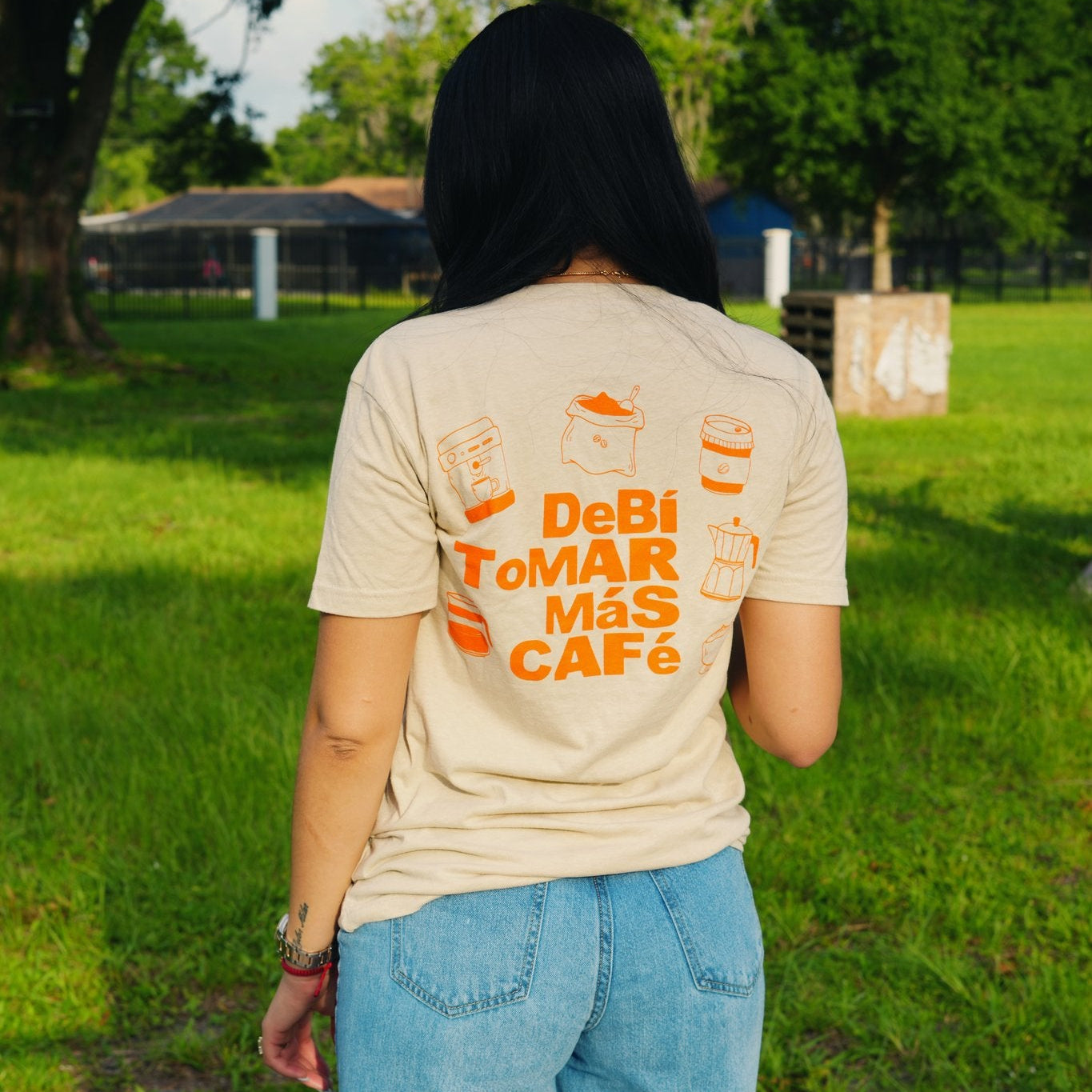 A person standing outdoors on a grassy lawn, viewed from behind, wearing a beige graphic T-shirt with orange text reading 'DeBi ToMar Más CAFE' and blue jeans.