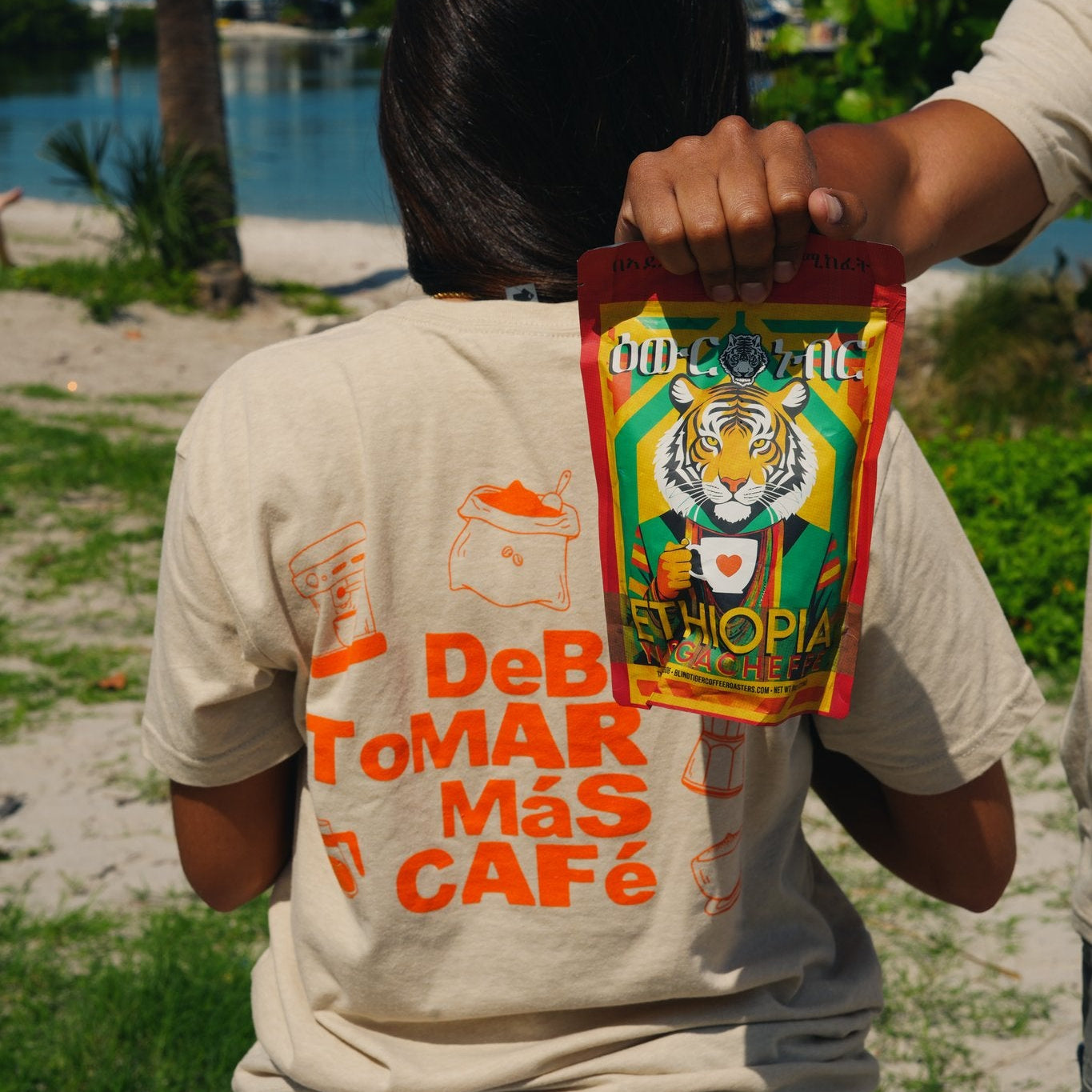 A person standing outdoors by the water holds up a colorful pouch of coffee behind their back. The person is wearing a beige t-shirt with orange and red text that reads 'DeB ToMAR Más CAFé' on the back.