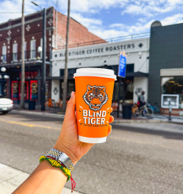 Person holding a 'Blind Tiger' coffee cup on a city street with buildings in the background.