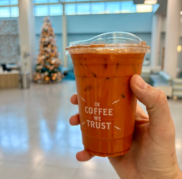 Hand holding a cup of iced coffee with 'In Coffee We Trust' text in an indoor setting.